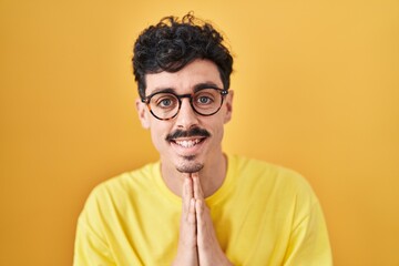 Hispanic man wearing glasses standing over yellow background praying with hands together asking for forgiveness smiling confident.