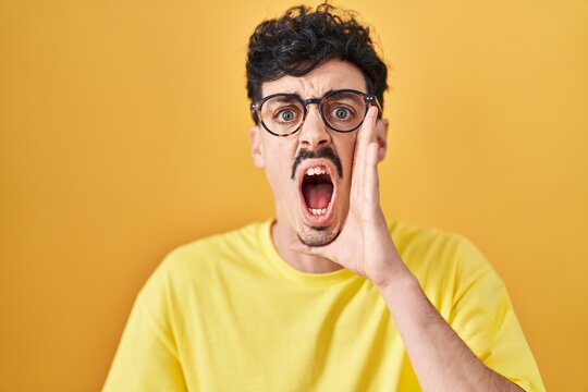 Hispanic man wearing glasses standing over yellow background shouting and screaming loud to side with hand on mouth. communication concept.