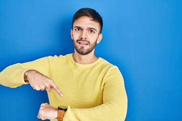 Hispanic man standing over blue background in hurry pointing to watch time, impatience, upset and angry for deadline delay