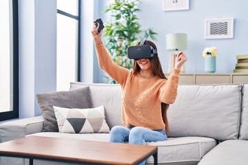 Young beautiful hispanic woman playing video game using virtual reality glasses and joystick at home