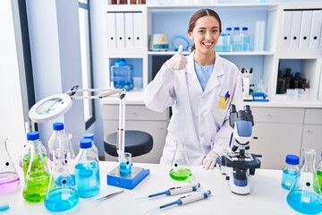 Young brunette woman working at scientist laboratory smiling happy and positive, thumb up doing excellent and approval sign