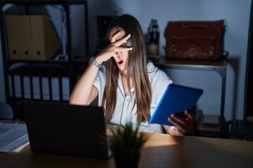 Young brunette woman working at the office at night peeking in shock covering face and eyes with hand, looking through fingers with embarrassed expression.