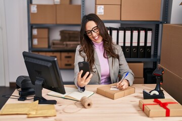 Young hispanic woman ecommerce business worker using smartphone writing on notebook at office