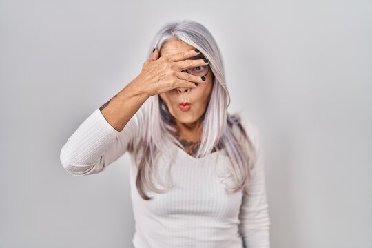 Middle age woman with grey hair standing over white background peeking in shock covering face and eyes with hand, looking through fingers with embarrassed expression.