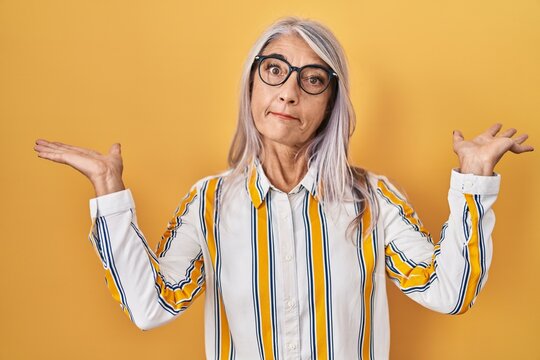 Middle age woman with grey hair standing over yellow background wearing glasses clueless and confused expression with arms and hands raised. doubt concept.