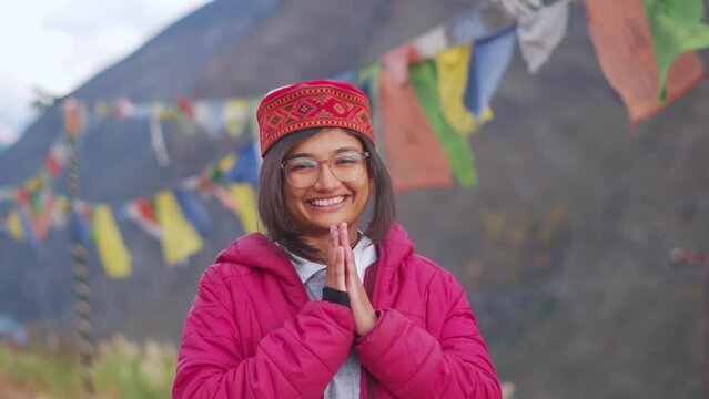 Portrait of happy smiling Indian woman wearing Himachali Cap standing at Himalayan mountains in background and doing Namaste. Positive emotion in a young Female. Indian tradition of greeting people.