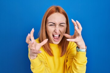 Young woman standing over blue background shouting frustrated with rage, hands trying to strangle, yelling mad