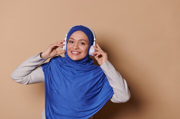 Isolated portrait on beige background of a charming modern Arab Muslim woman,with head covered in a blue hijab, listening to to a new sound track on wireless headphones and smiling looking at camera