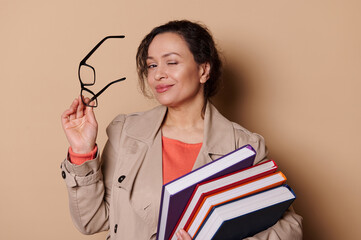 Close-up portrait of a multi-ethnic elegant charming woman in casual wear, winking and smiling looking at camera, holding stylish black spectacles and hardcover books, on beige isolated background