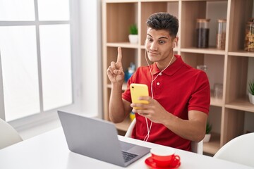 Young hispanic man using laptop and doing video call with smartphone smiling with an idea or question pointing finger with happy face, number one