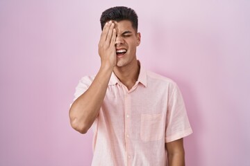 Young hispanic man standing over pink background yawning tired covering half face, eye and mouth with hand. face hurts in pain.