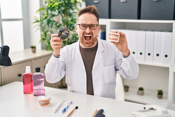 Middle age caucasian dentist man holding denture and doughnuts angry and mad screaming frustrated and furious, shouting with anger. rage and aggressive concept.