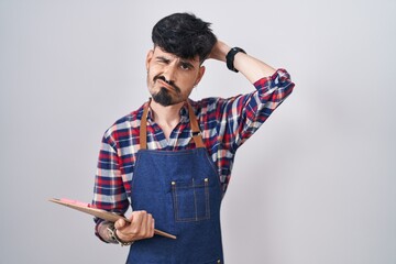 Young hispanic man with beard wearing waiter apron holding clipboard confuse and wondering about...