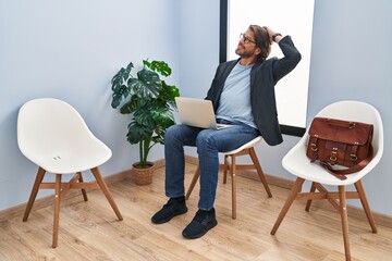 Handsome middle age man sitting at waiting room working with laptop smiling confident touching hair with hand up gesture, posing attractive and fashionable