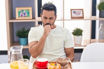 Hispanic man with beard eating breakfast feeling unwell and coughing as symptom for cold or...