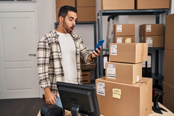 Young hispanic man with beard working at small business ecommerce with smartphone scared and amazed...