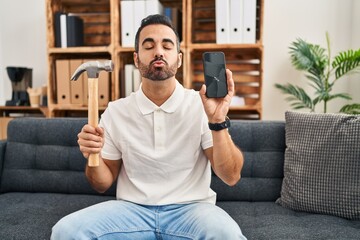 Young hispanic man with beard holding hammer and broken smartphone showing cracked screen looking...