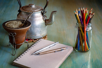 Cuaderno con lapiceras sobre la mesa, haciendo tareas en la mañana con luz natural suave, pava y mate de fondo, frasco de vidrio con lápices de colores y mesa vintage © Daniel