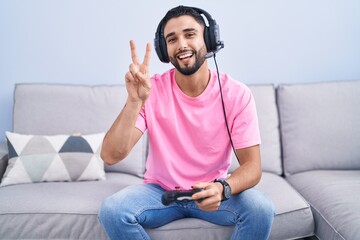 Hispanic young man playing video game holding controller sitting on the sofa smiling with happy face winking at the camera doing victory sign with fingers. number two.