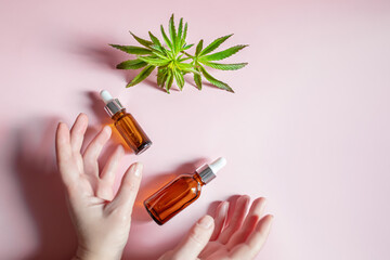Women's hands holding cosmetic bottles on a pink background. Fresh hemp leaves to make elexir for cosmetics