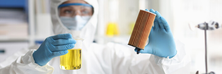 Scientist in protective suit holds car filter cartridge in laboratory and oil - obrazy, fototapety, plakaty