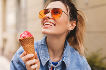 Woman with high ponytail hold delicious pink ice cream in waffle cone outdoors. Girl wear orange...