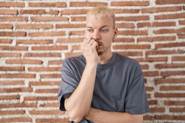 Young caucasian man standing over bricks wall looking stressed and nervous with hands on mouth biting nails. anxiety problem.