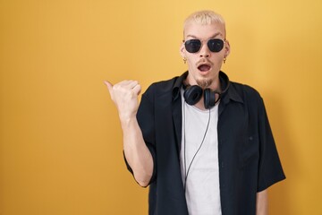 Young caucasian man wearing sunglasses standing over yellow background surprised pointing with hand finger to the side, open mouth amazed expression.