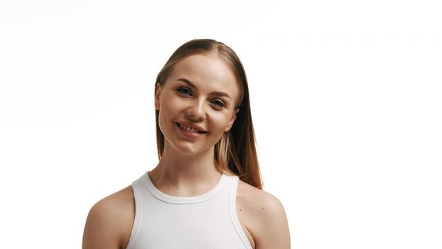 Fit attractive european blonde skin care woman touching her bare shoulder and turning her head to the camera looking at it and smiling broadly against a white background.