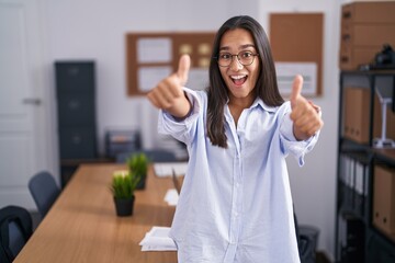 Young hispanic woman at the office approving doing positive gesture with hand, thumbs up smiling...