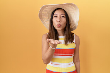 Middle age chinese woman wearing summer hat over yellow background looking at the camera blowing a kiss with hand on air being lovely and sexy. love expression.