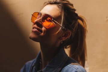 Attractive woman with high ponytail standing near beige wall on the street of old city. Girl joy of sun light, wear orange sunglasses look at sun. Shadow and light of sunny summer, spring or fall day