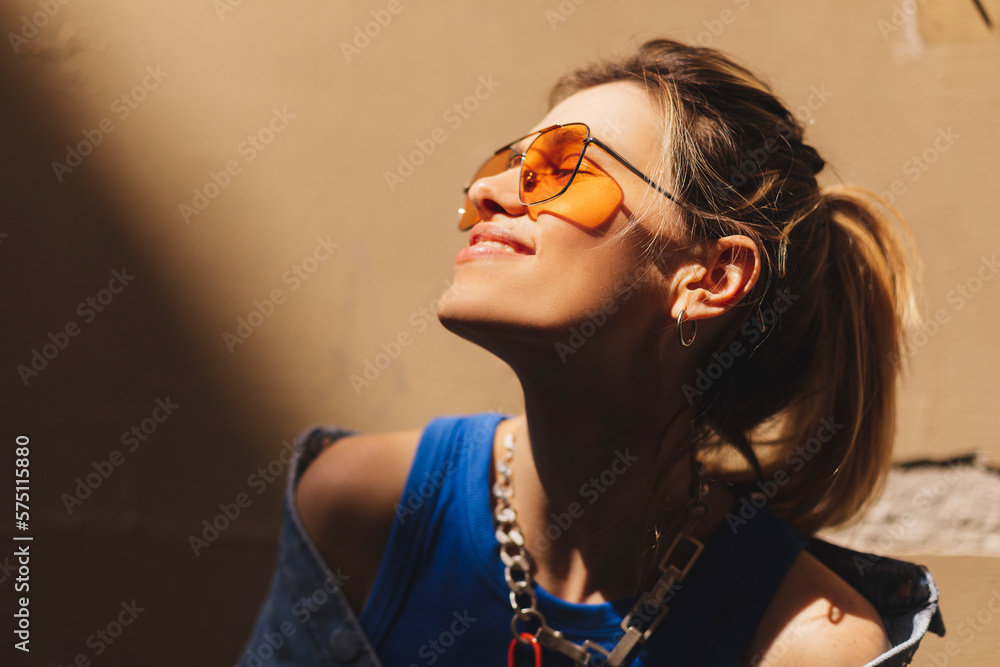 Sticker Attractive woman with high ponytail standing near beige wall on the street of old city. Girl joy of sun light, wear orange sunglasses look at sun. Shadow and light of sunny summer, spring or fall day