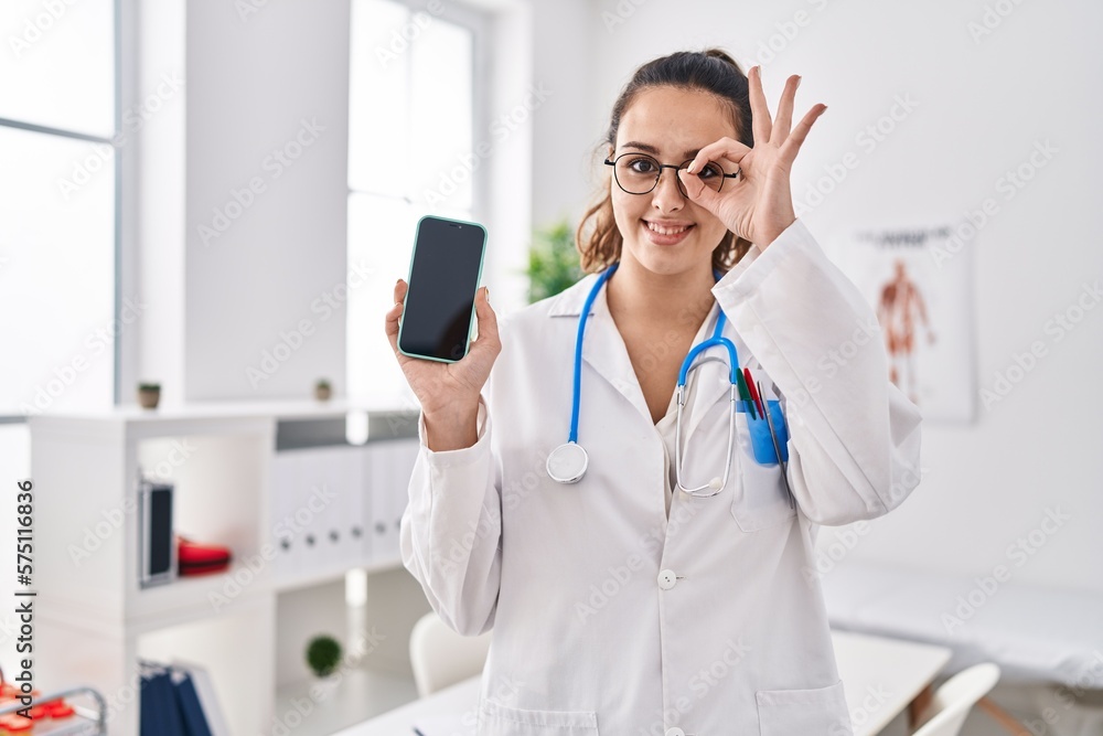 Sticker young hispanic doctor woman holding smartphone showing screen smiling happy doing ok sign with hand 