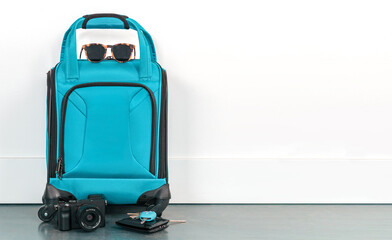 Turquoise blue travel bag with sunglasses, camera, wallet, and keys against a plain white wall. Ready for a vacation holiday.