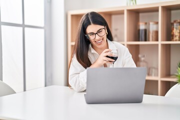 Young hispanic woman using laptop drinking wine at home