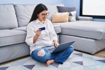 Young hispanic woman using laptop and credit card sitting on sofa at home