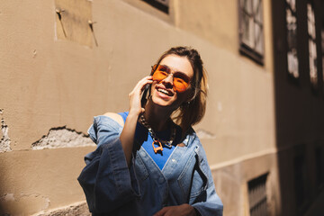 Beautiful young woman wearing denim shirt, blue top and orange sunglasses walking outdoors, talking on mobile phone. Portrait of stylish smiling woman ponytail calling on mobile phone on old street.