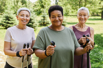 Waist up portrait of three sportive senior women smiling at camera holding walking poles outdoors