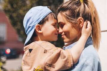 Happy mom holding son boy in arms, hugging kid with noses touch, smiling, enjoying warm moment. Younger mum carrying kid and dancing on the street. Motherhood, adoption, family concept.