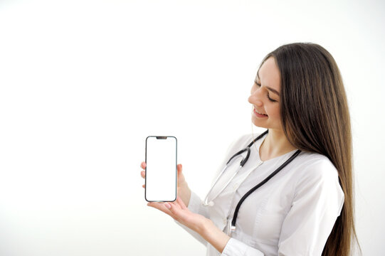 Woman doctor or nurse wearing a lab coat and stethoscope pointing to a blank mobile phone with a pleased smile close up over a grey background. High quality photo