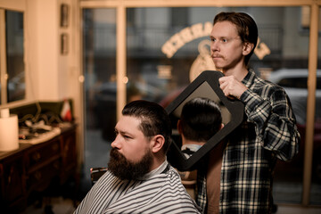 side view of bearded man in a barbershop and barber shows hairstyle with mirror