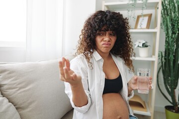 Fototapeta na wymiar Pregnant woman smile blogger taking vitamins and medicine pills with glass of water sitting on sofa at home freelancer in last month of pregnancy lifestyle before childbirth