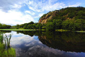 Fototapeta premium Lake and mountain range on a sunny summer day. The blue sky with clouds is reflected on the water surface. Beautiful summer landscape with a lake
