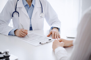 Fototapeta premium Doctor and patient sitting opposite each other at the desk in clinic. The focus is on female physician's hands filling up the medication history record form or checklist, close up. Medicine concept