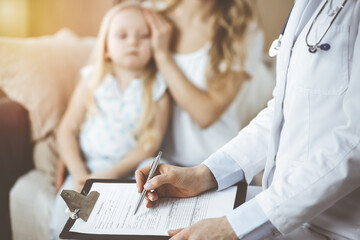Fototapeta premium Doctor and patient. Pediatrician using clipboard while examining little girl with her mother at home. Sick and unhappy child at medical exam