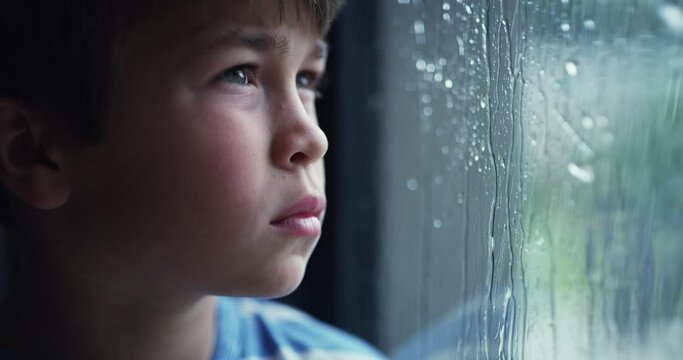A sad little boy depressed by bad weather, sitting inside watching rain through a window. Disappointed child unhappy, bored, lonely and frustrated by failed plans. Kid stuck indoors due to a storm
