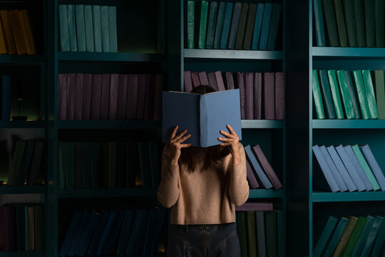 A student in the library, preparing for exams, studying at the university. A young woman with a blue open book on the background of bookshelves.