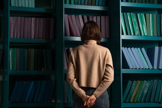 Apathy And Frustration. Cognitive Overload From An Overabundance Of News And Information Noise, Concept. Back View Of A Woman Standing With Her Face Buried In Bookshelves.