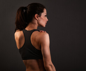 Fototapeta na wymiar Serious female sporty muscular with ponytail doing stretching workout the shoulders, blades and arms in sport bra, standing on dark grey background with empty copy space. Back view.